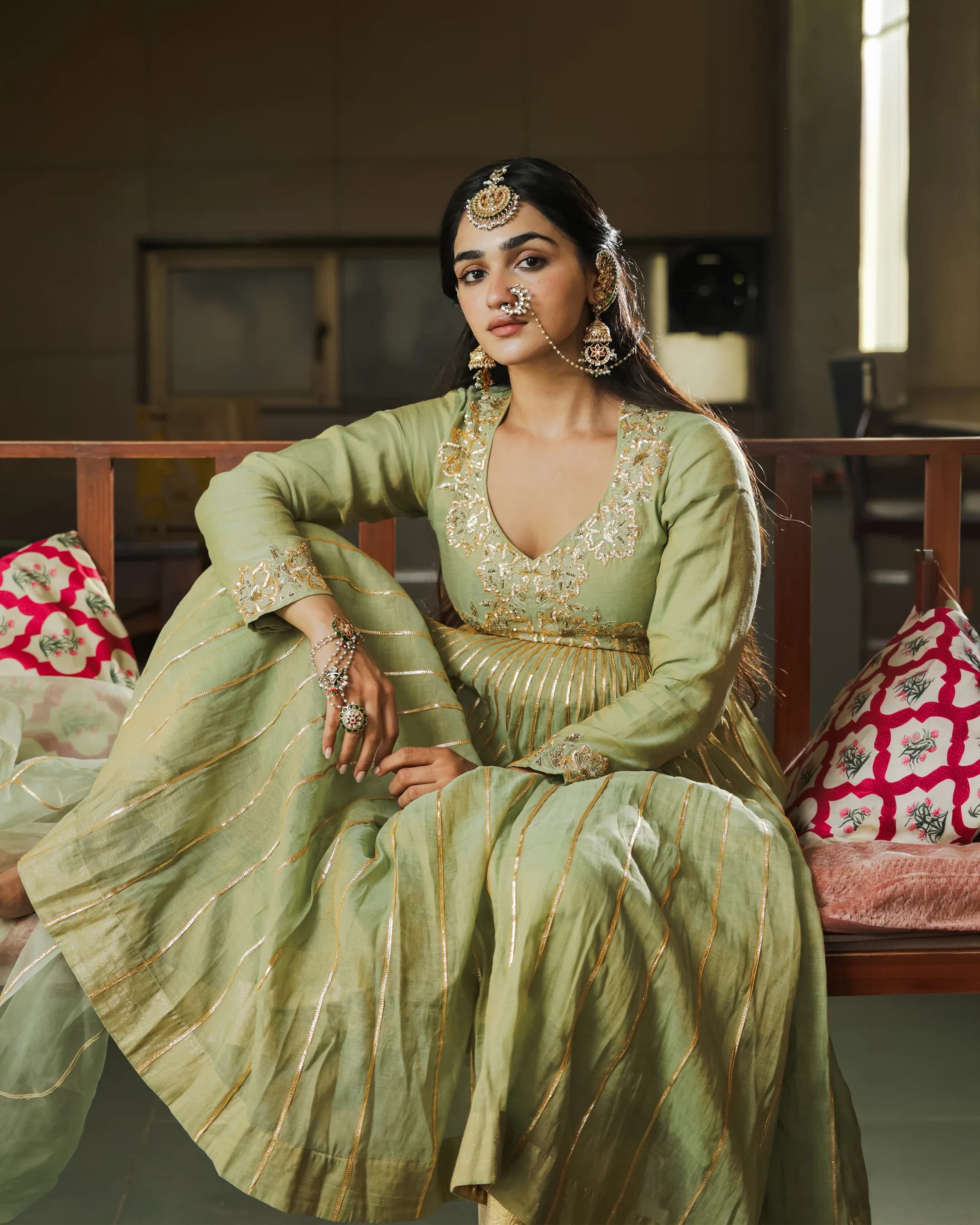 Model in a pistachio green anarkali with gold gota stripes, seated on a wooden daybed with printed cushions