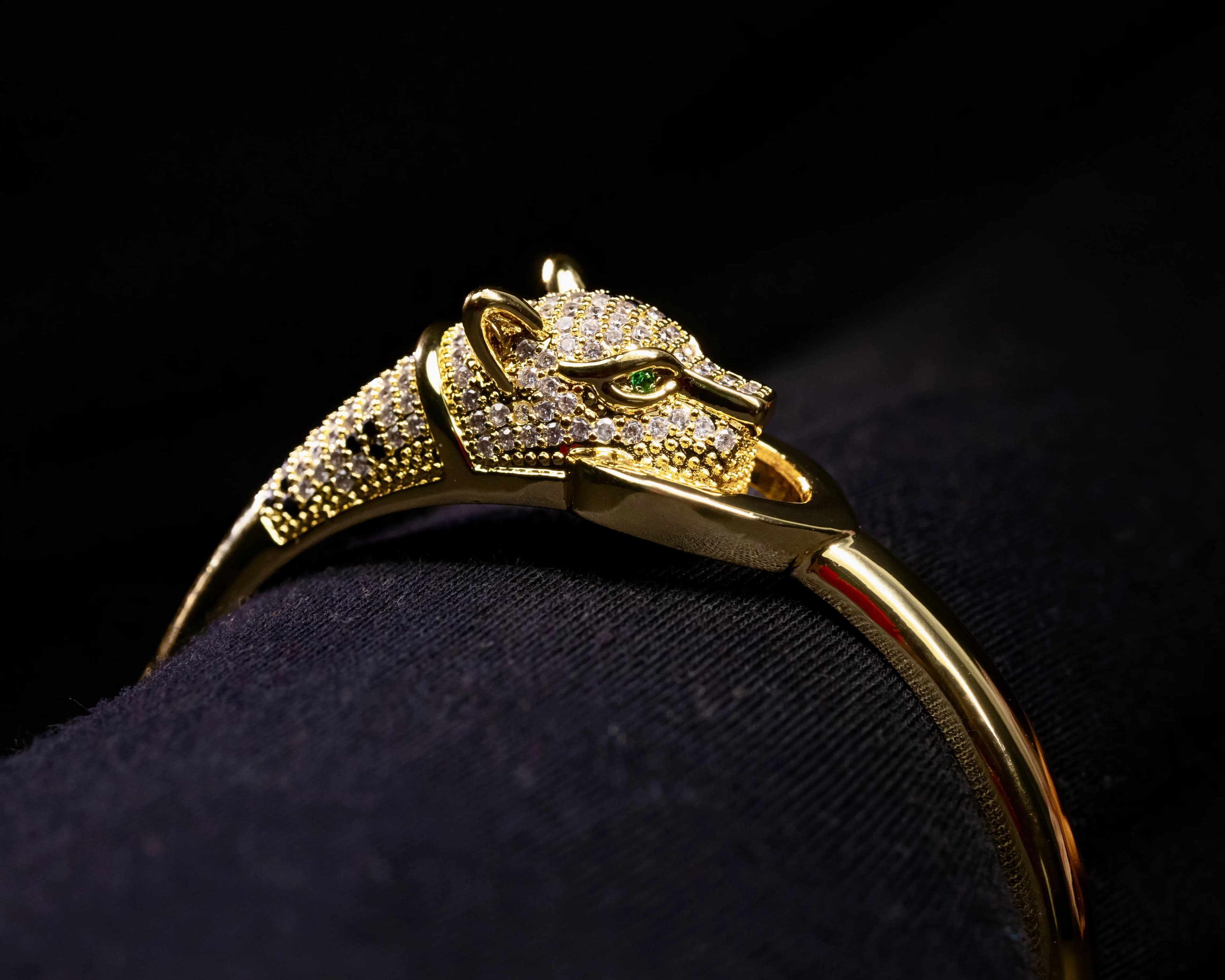 Close-up of a gold panther-head bangle with pave stones and a green eye on dark fabric