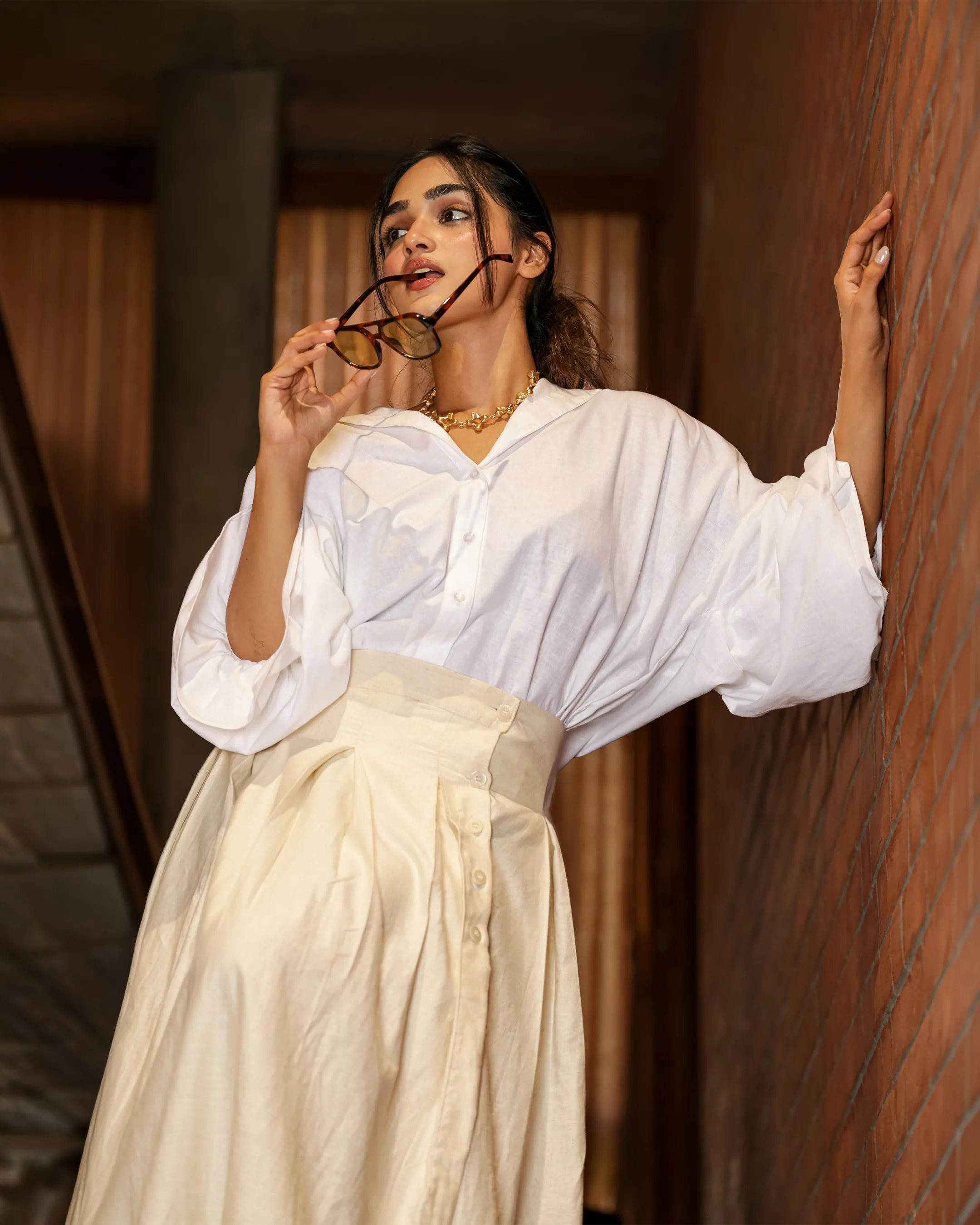 Model in an oversized white shirt and pleated high-waist cream skirt, leaning on a brick wall with sunglasses