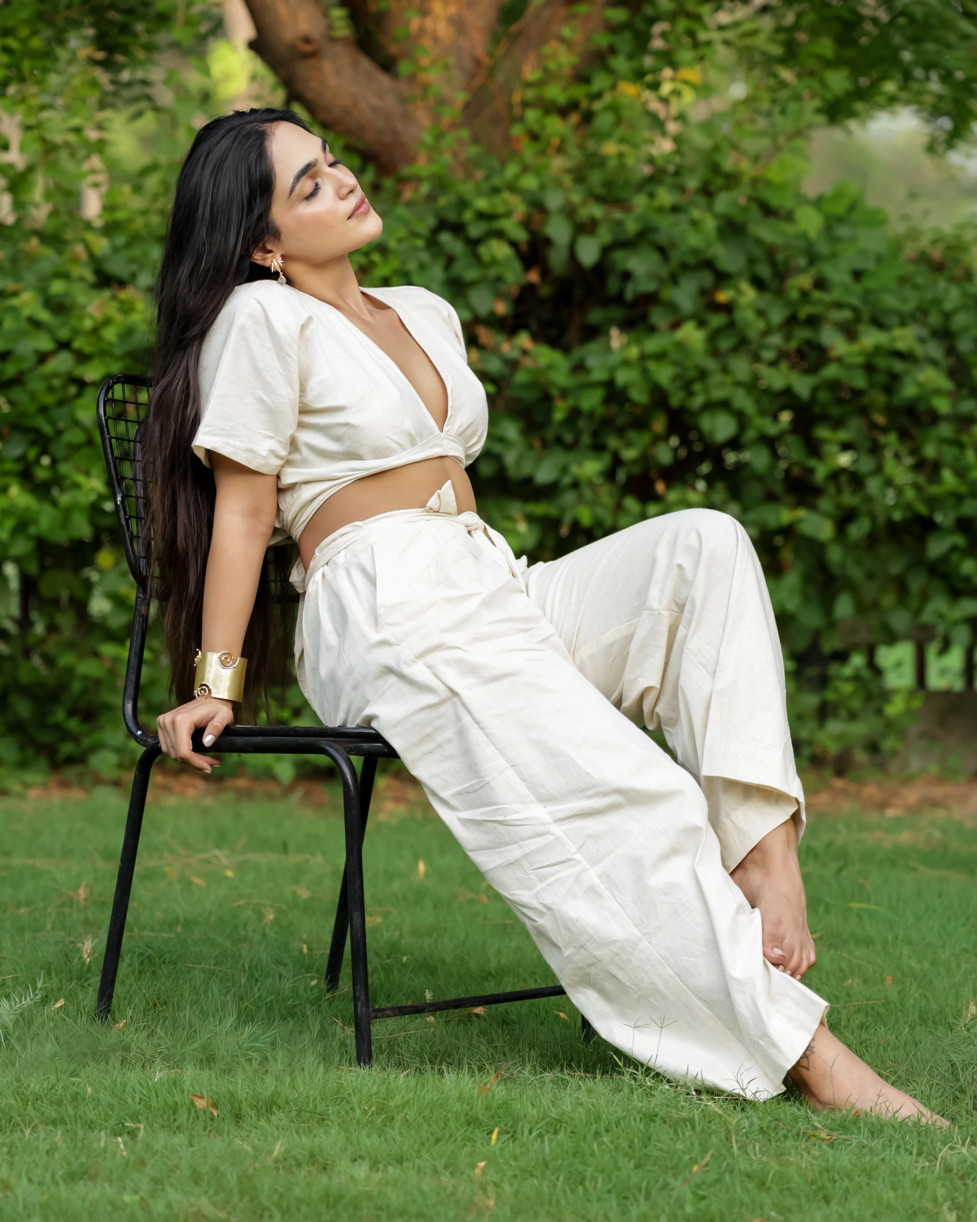 Model in a cream wrap crop top and wide-leg pants, reclining on a black chair on a lawn