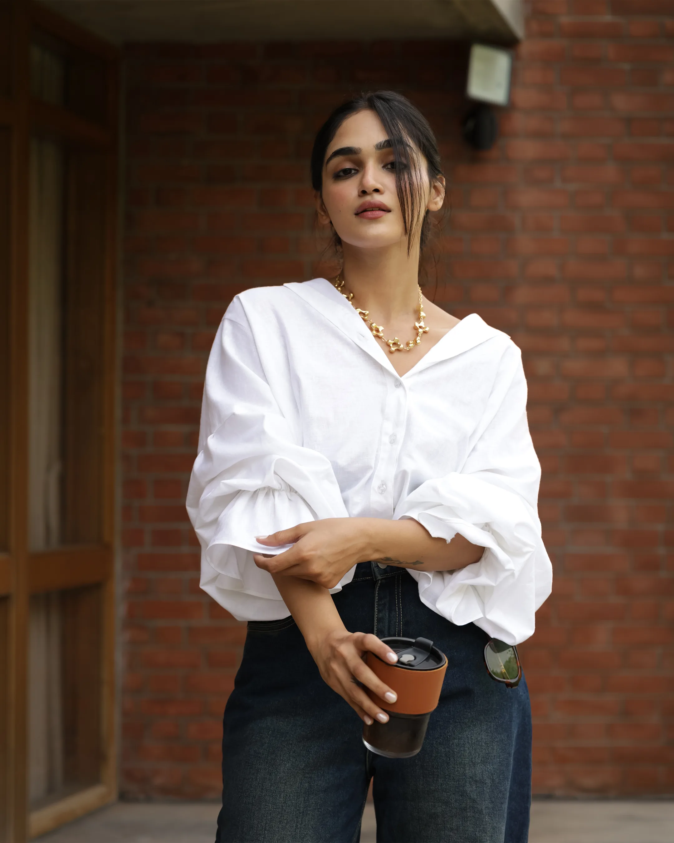 Model in a white balloon-sleeve shirt and denim, holding a coffee cup against a brick wall