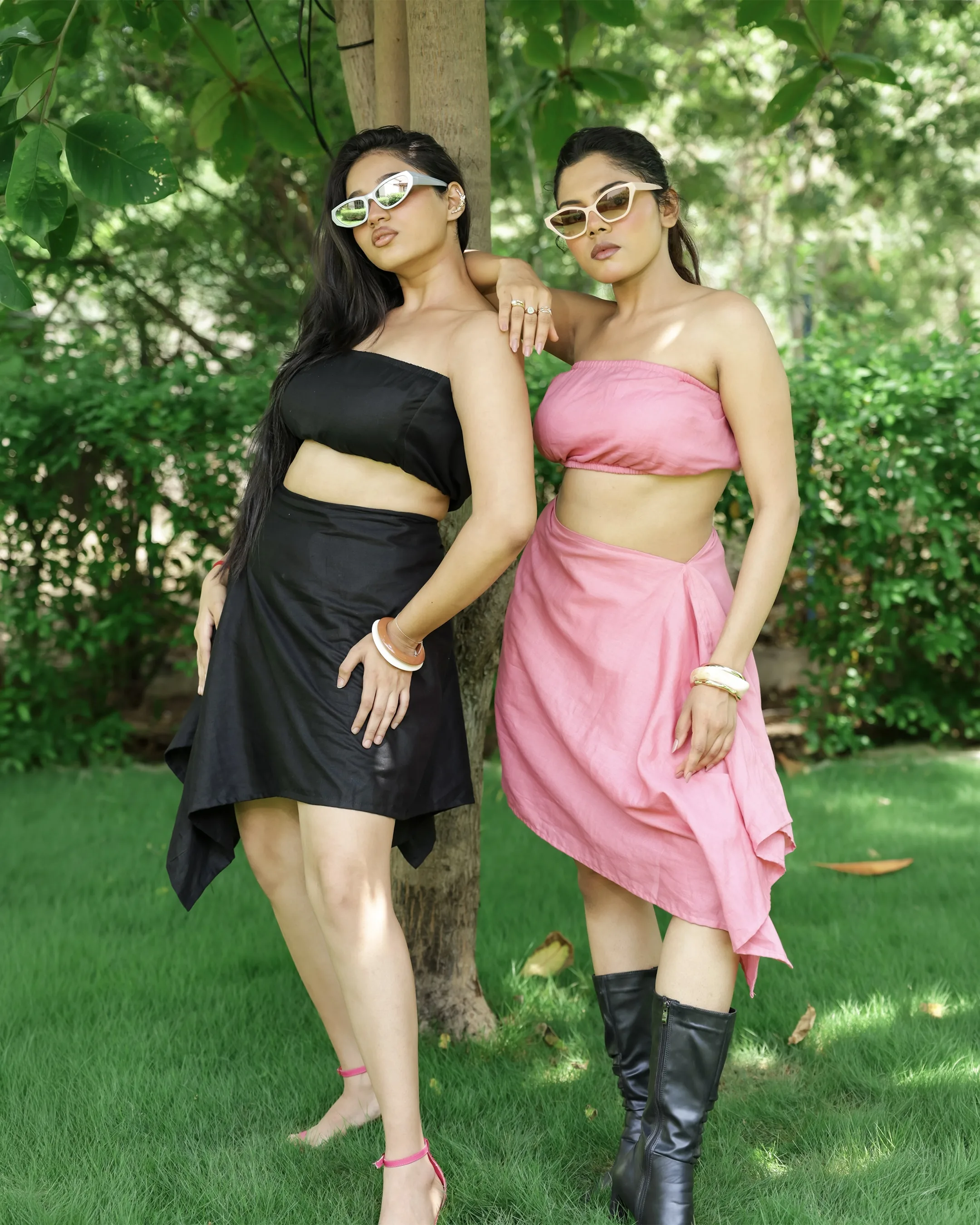 Two models in matching strapless tops and asymmetric skirts, one black and one pink, under a tree