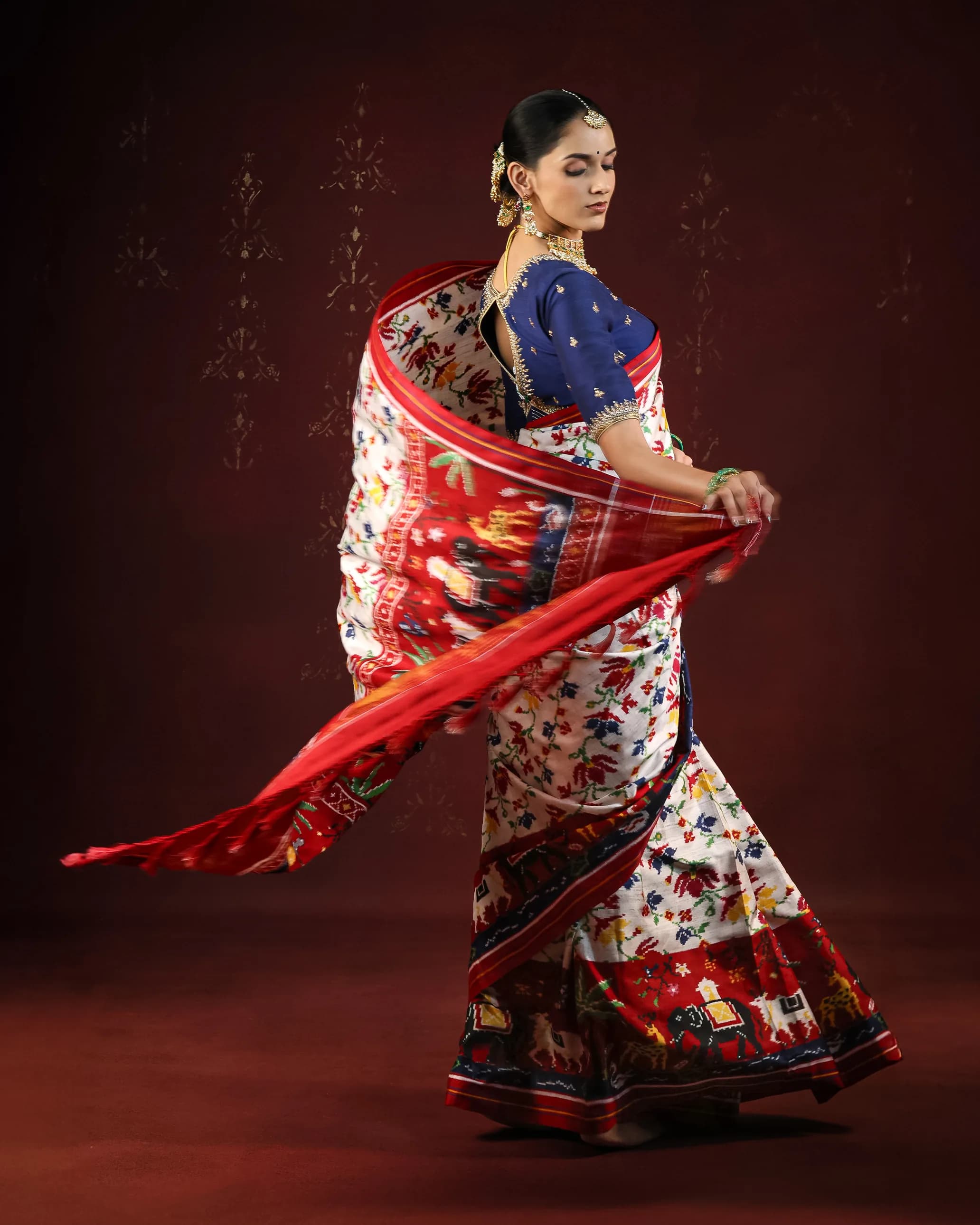 Model twirling in a cream and red patola saree with an elephant border and navy blouse, against a deep maroon backdrop