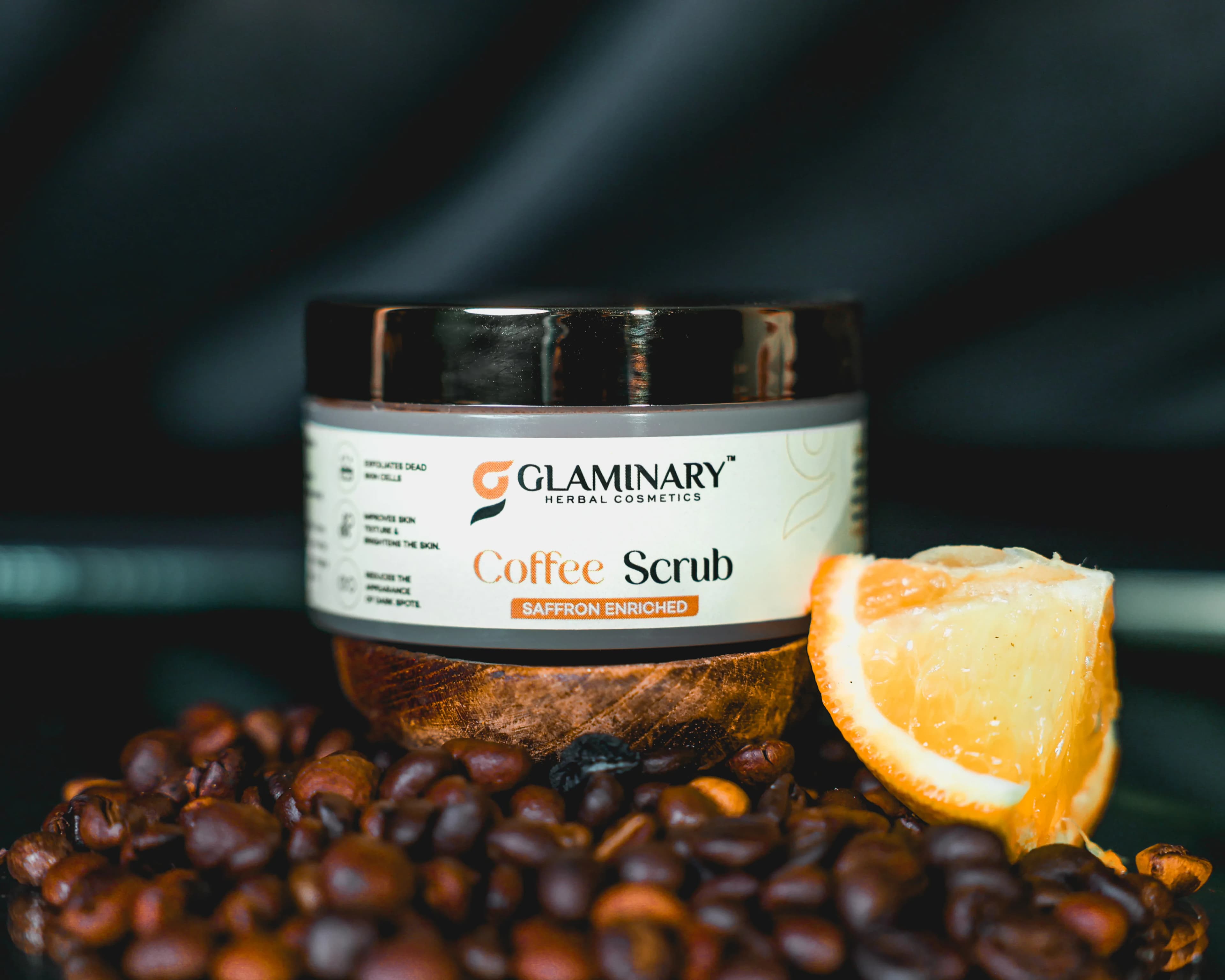 Glaminary Coffee Scrub jar on a wooden bowl of coffee beans with an orange slice, dark backdrop