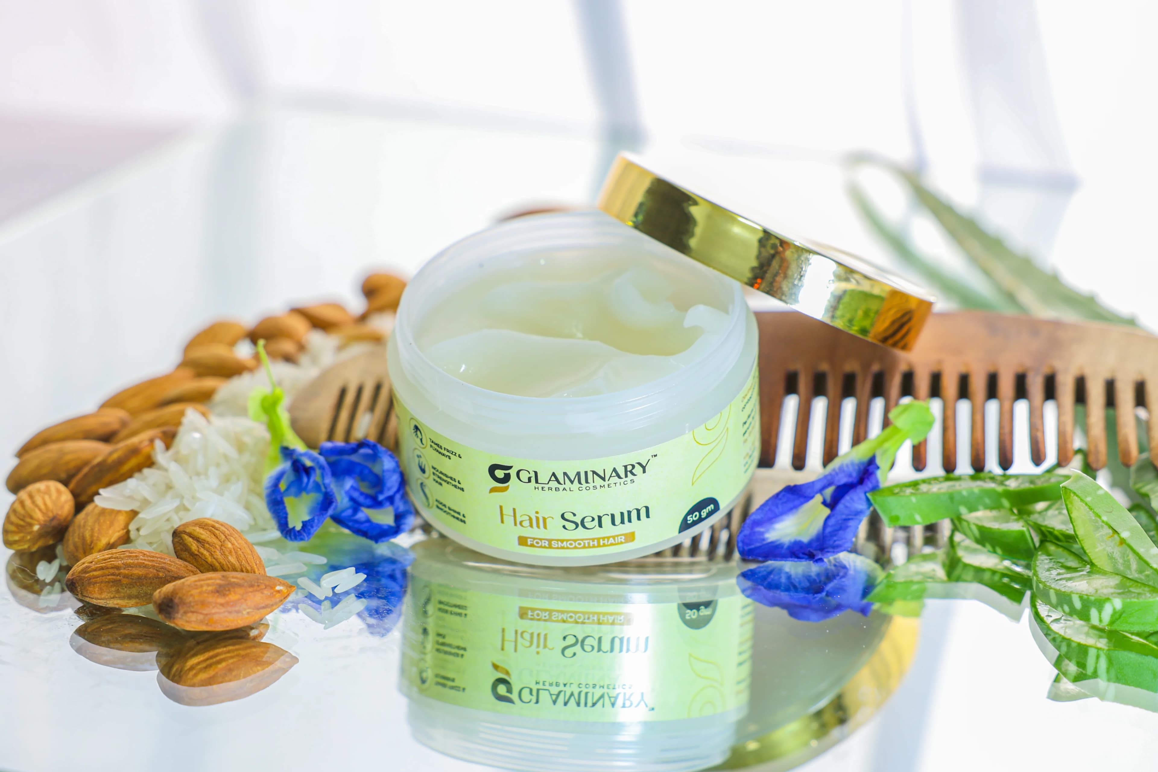 Open jar of Glaminary Hair Serum with almonds, rice, blue pea flowers, aloe and a wooden comb on glass