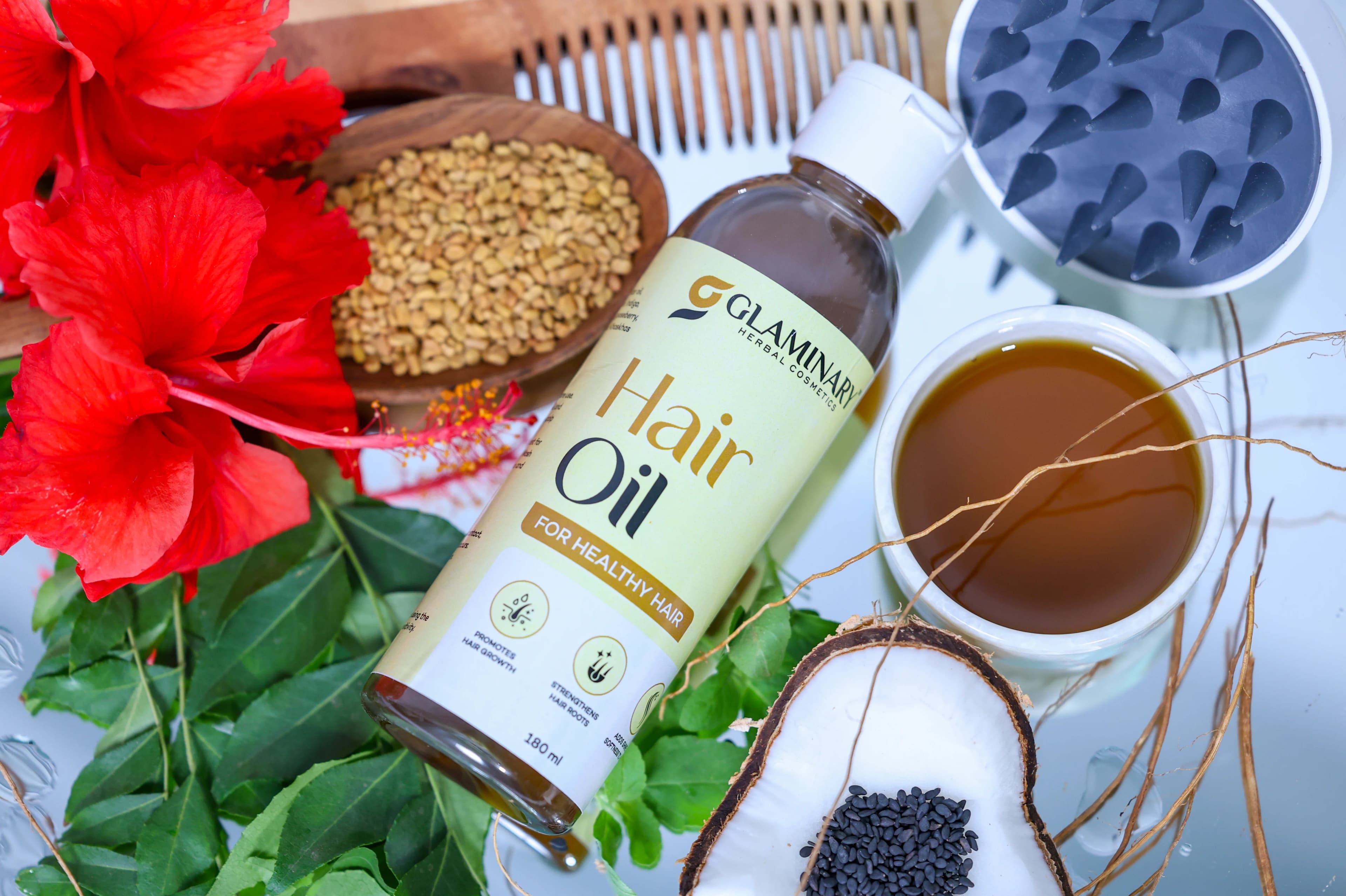 Glaminary Hair Oil bottle laid among hibiscus flowers, fenugreek seeds, curry leaves and coconut