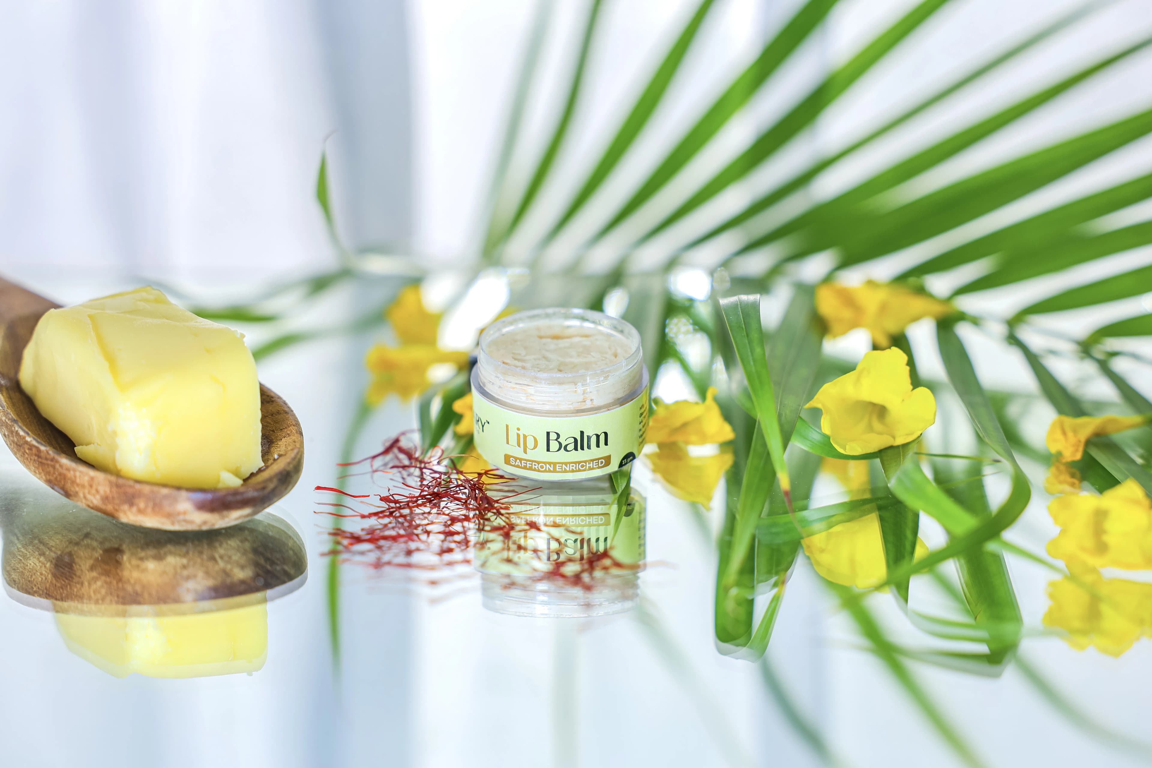Open Glaminary Lip Balm with saffron strands, ghee on a wooden spoon and yellow flowers