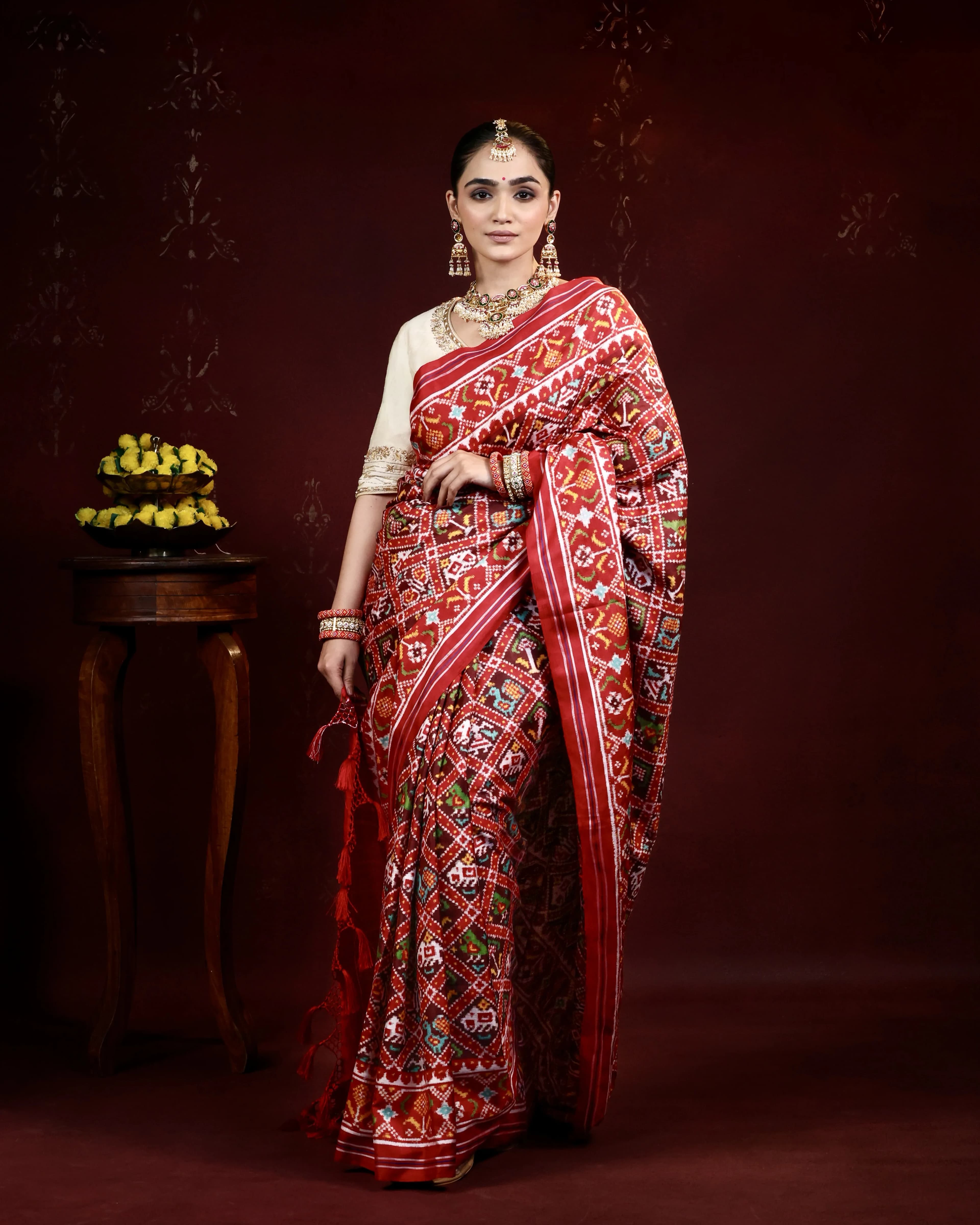 Model in a red geometric patola saree with a cream blouse, beside a table of marigolds