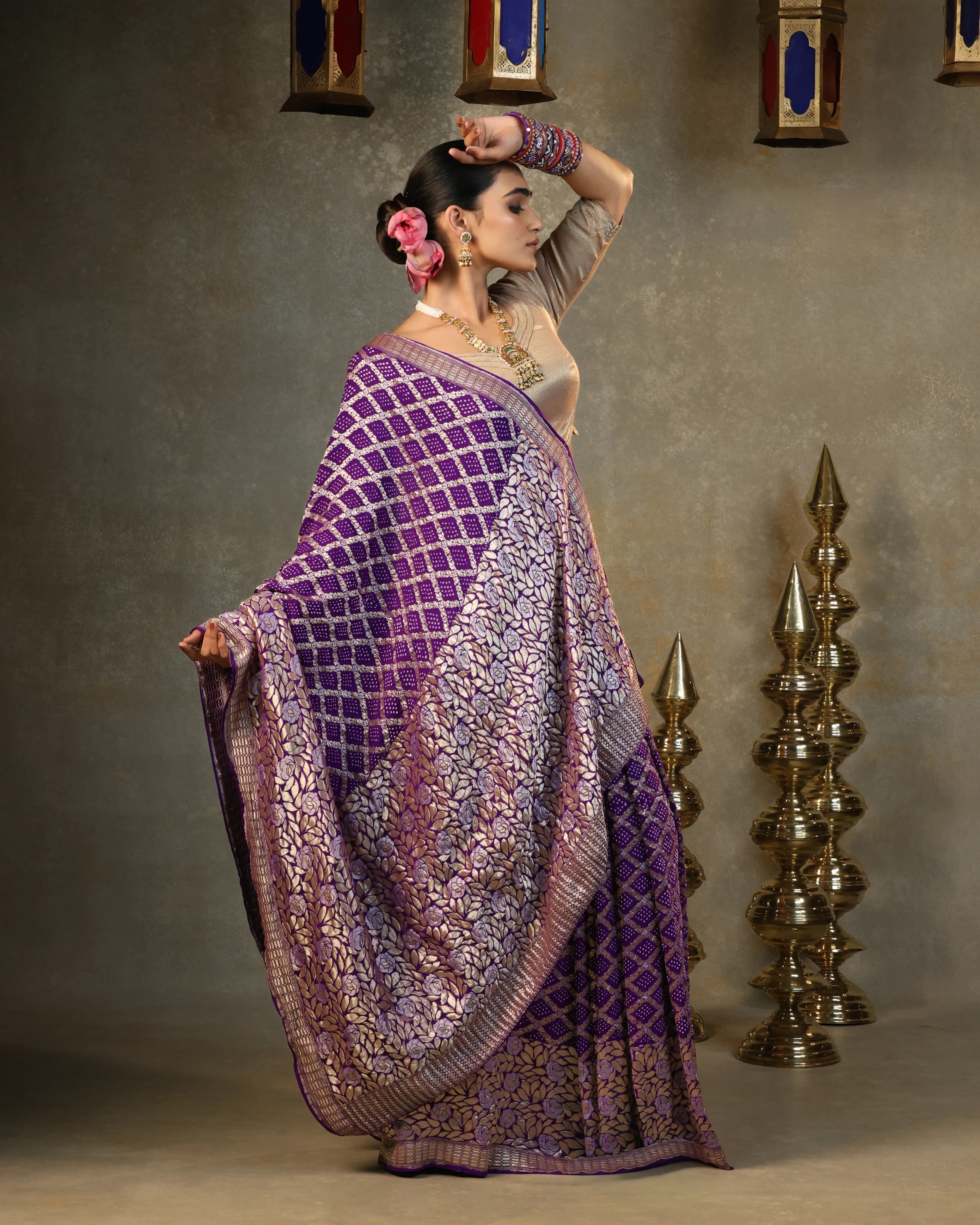 Model in a purple bandhani saree with gold zari florals, arm raised, beside brass lamps
