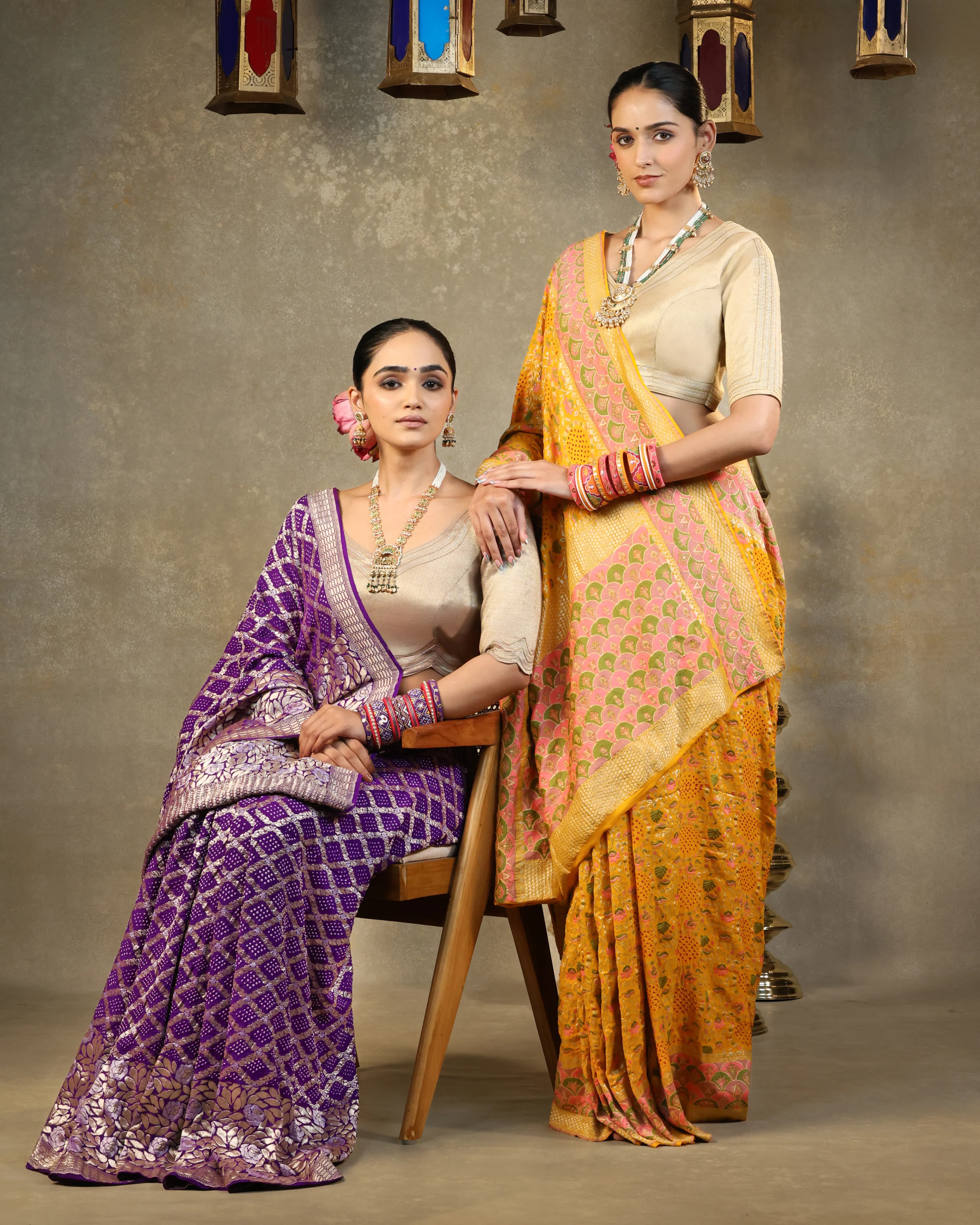 Two models in purple and yellow bandhani sarees under hanging lanterns